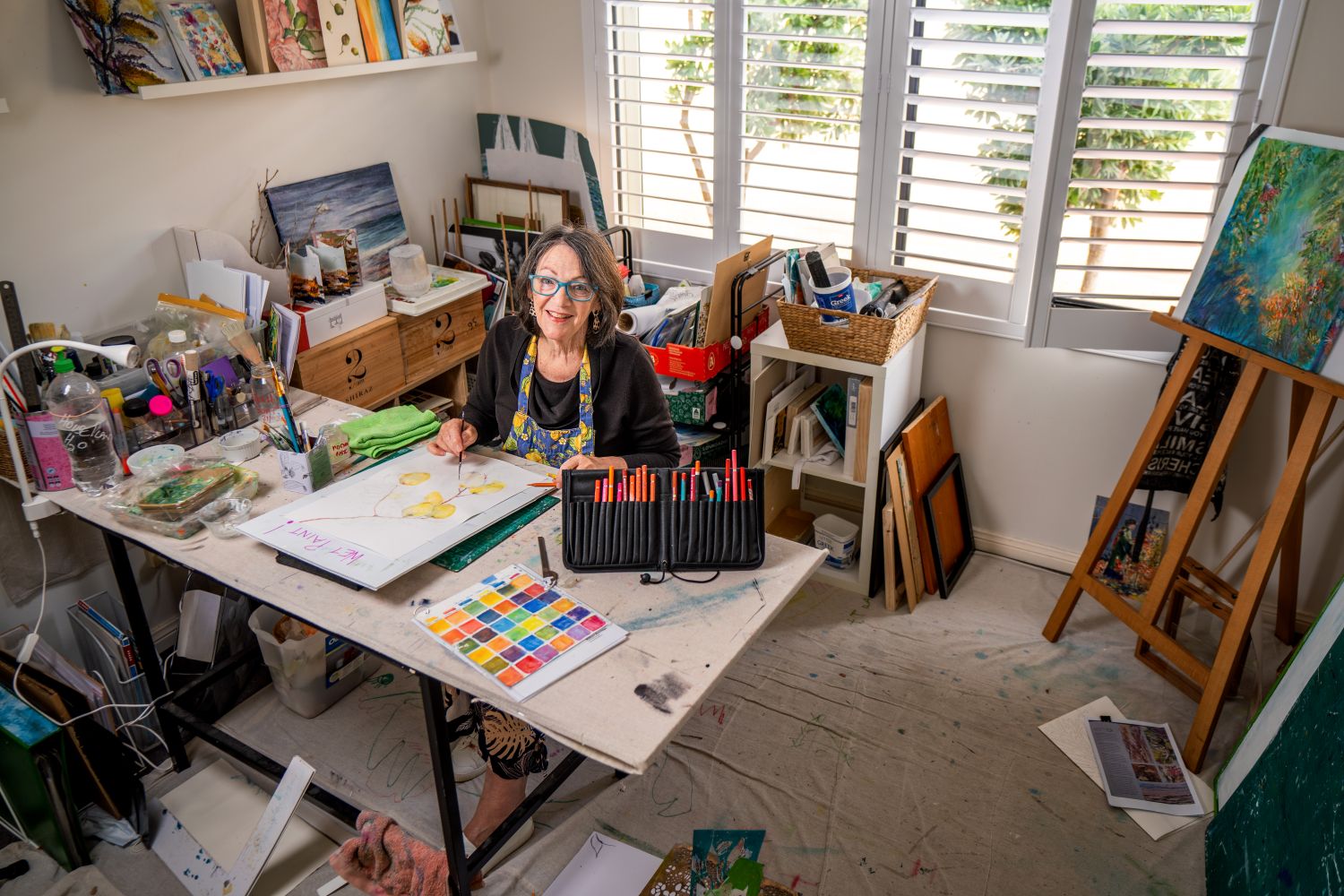A woman sits at a table in a bright, colourful art studio, surrounded by pencils, paper and art supplies, smiling